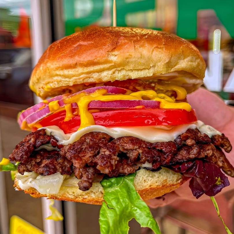 Close-up of a Peri Peri Original burger with two patties, cheese, tomato, red onion, and mustard.