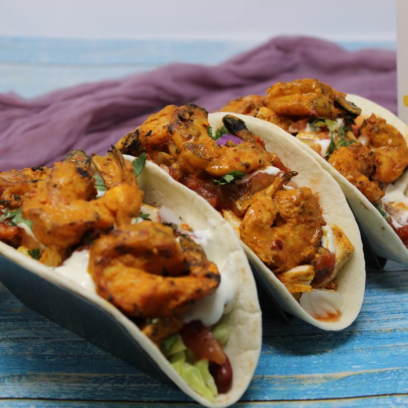 Three Peri Peri Original shrimp tacos with sauce and greens on a blue wooden table.